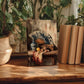 a wooden table with a framed painting of a fan and various objects, including a vase, a fruit bowl, and some books, placed on it. The table is surrounded by potted plants, creating a cozy and inviting atmosphere.